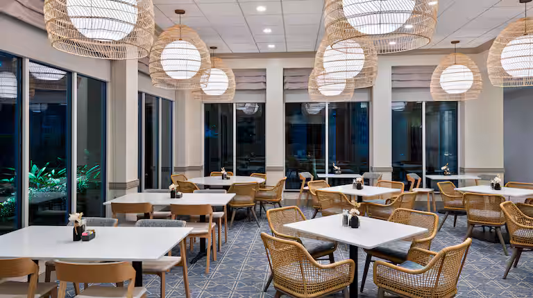 A modern, empty restaurant with light wood chairs, white tables, and large round wicker pendant lights. Floor-to-ceiling windows line the walls, and patterned carpet covers the floor. The atmosphere is clean and inviting.