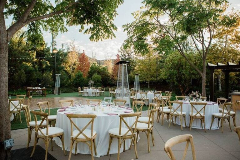 Outdoor event setup with round tables covered in white cloths, wooden chairs, and neatly arranged tableware. The area is surrounded by trees and greenery, with patio heaters and string lights overhead under a clear sky.