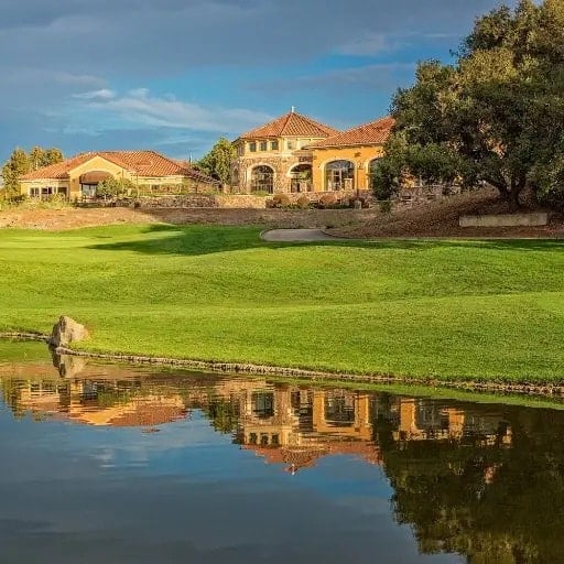 A large, elegant house with arched windows and a red-tiled roof sits beside a golf course, reflected in a calm pond under a partly cloudy blue sky.