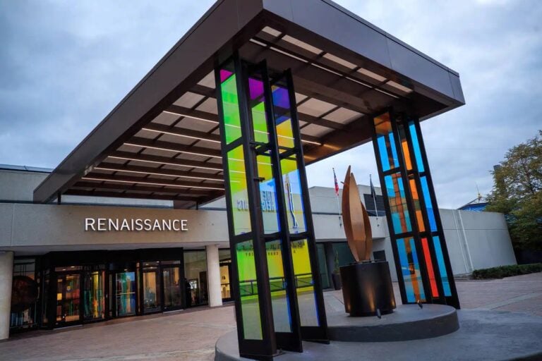 Entrance to a modern building labeled Renaissance, featuring colorful glass panels, a geometric sculpture, and large windows under a covered walkway. Trees and cloudy sky are visible in the background.