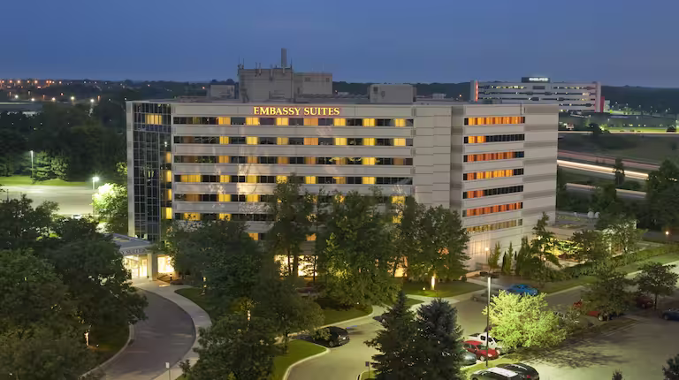 A multi-story Embassy Suites hotel is illuminated at dusk, with warm lights glowing from many windows. The building is surrounded by trees and a curved driveway, with other distant buildings and roads visible in the background.