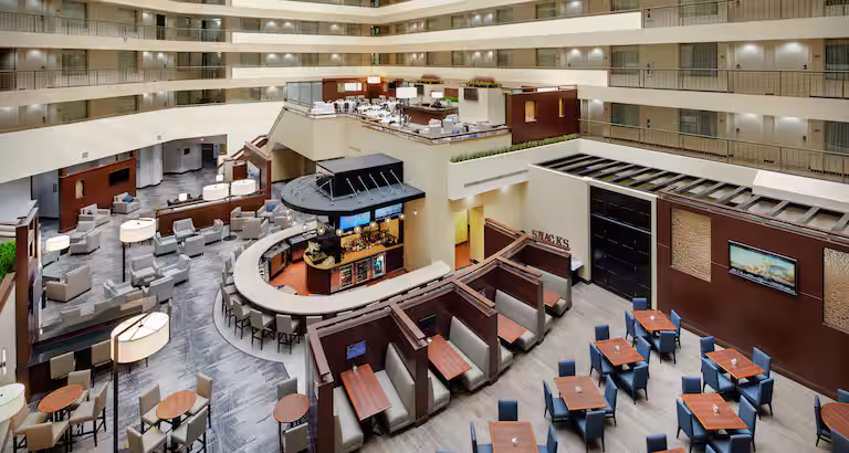 Spacious hotel atrium with multiple floors and balcony railings overlooking a modern dining area featuring booths, tables, and a central bar in neutral colors, with soft lighting and contemporary decor.