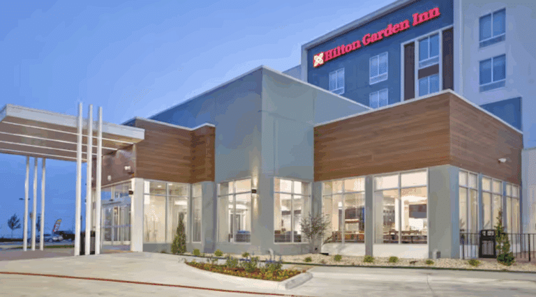 Modern exterior view of a Hilton Garden Inn hotel at dusk, featuring large glass windows, a covered driveway, landscaped greenery, and illuminated signage on the building.