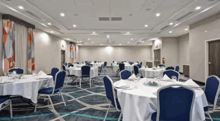 A spacious banquet hall with round tables covered in white tablecloths, blue chairs, neatly arranged place settings, and decorative curtains along the walls. The room is brightly lit with modern ceiling lights.