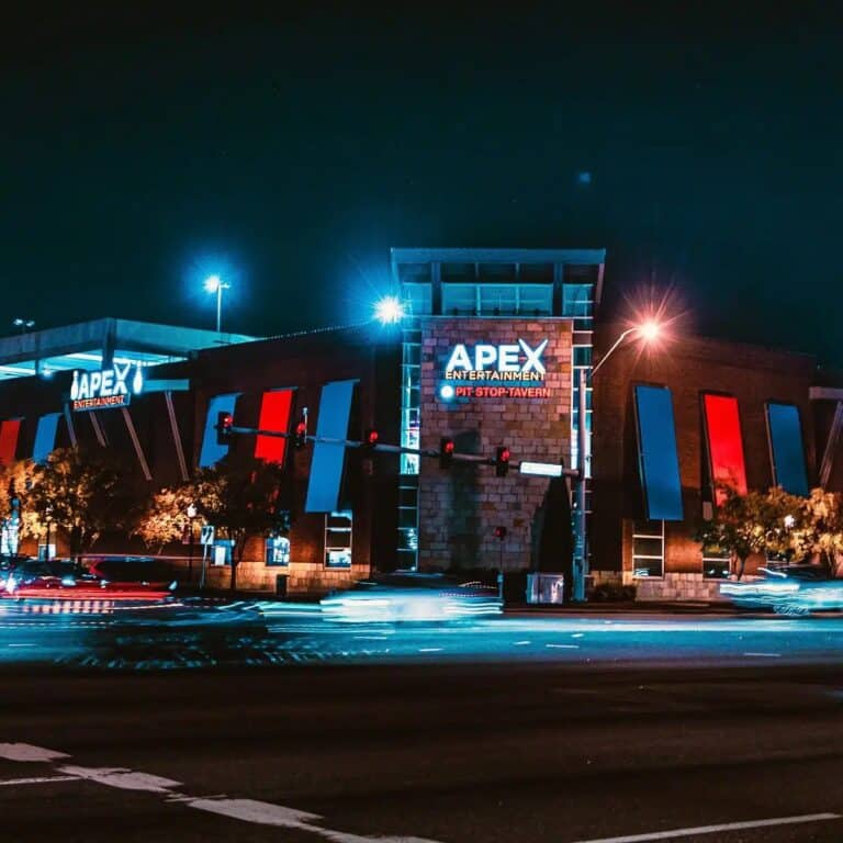 Apex Entertainment building at night, brightly lit with blue and red lights, cars passing by on the road in front, creating light trails.