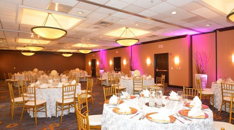 Hotel banquet hall set up for an event, with round tables covered in white cloths, gold chairs, elegant place settings, and purple accent lighting along the walls. Large ceiling lights illuminate the spacious room.
