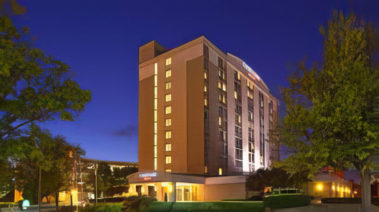 A tall, modern hotel building with warm exterior lights stands against a deep blue evening sky, surrounded by trees and greenery.