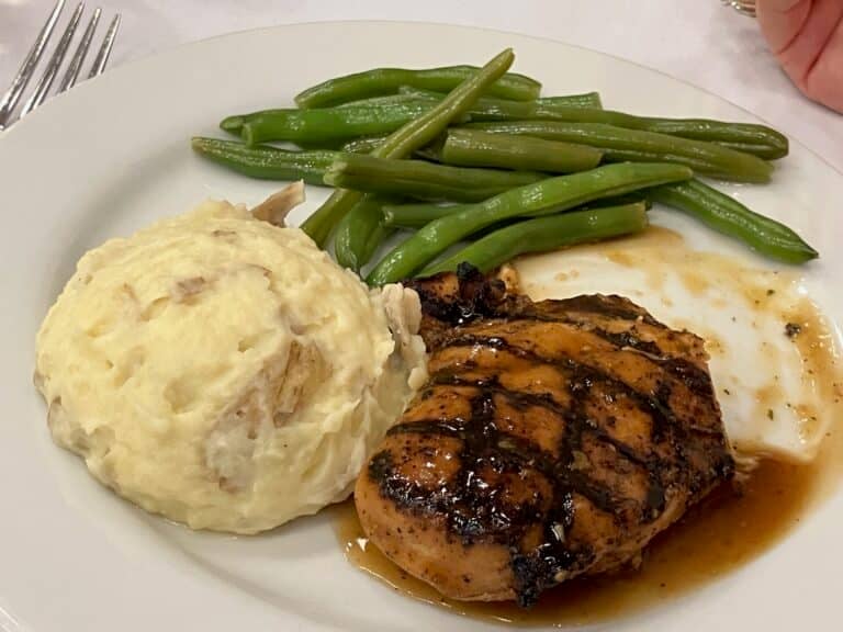 A plate with grilled chicken breast in sauce, a serving of mashed potatoes with skin, and steamed green beans.