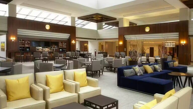 Spacious hotel lobby with modern seating, including cream and navy sofas with yellow pillows, small dark tables, and a bar area in the background under a large skylight ceiling.