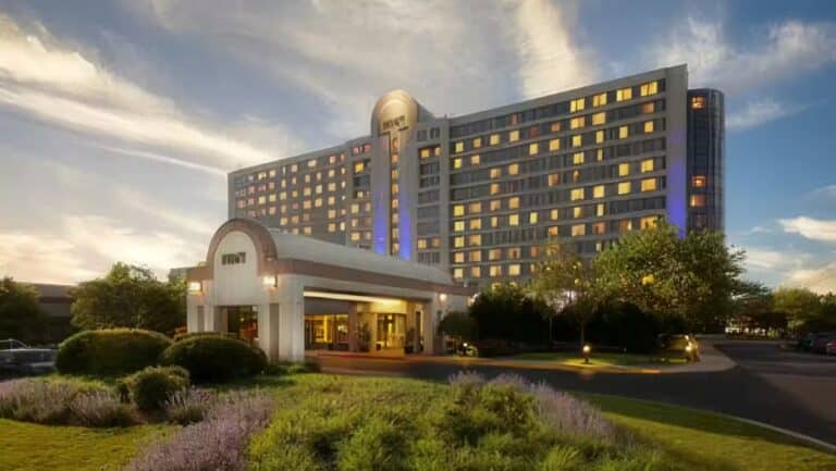 A large hotel building with numerous windows and a rounded entrance canopy, surrounded by green lawns, bushes, and trees, under a partly cloudy sky at either sunrise or sunset.