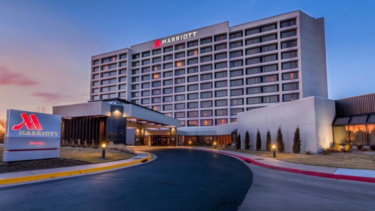 A large, modern Marriott hotel with multiple floors, large windows, and exterior lighting at dusk. The entrance driveway curves toward the main entrance, and a Marriott sign is visible in the foreground.