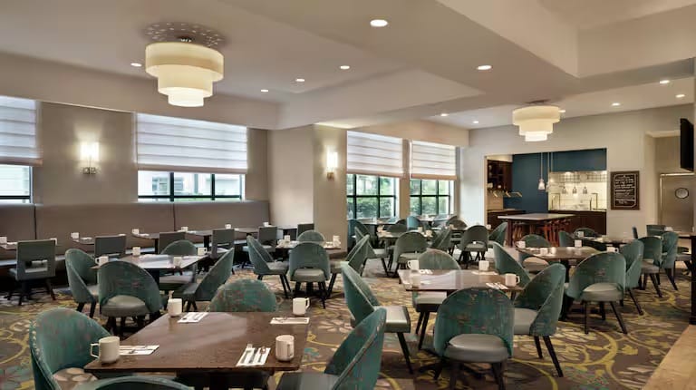 A modern, well-lit restaurant dining area with teal chairs, dark wood tables set with white mugs and cutlery, large windows with shades, and stylish ceiling lights. The room appears clean and inviting.