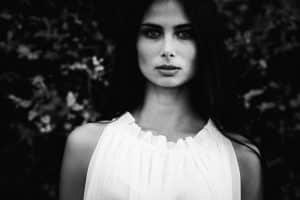 A black and white portrait captures a person with long dark hair and a serious expression, wearing a sleeveless white top. The backdrop of blurred dark foliage adds an element of mystery, offering variety without revealing any clear clue to the true mood behind the gaze.