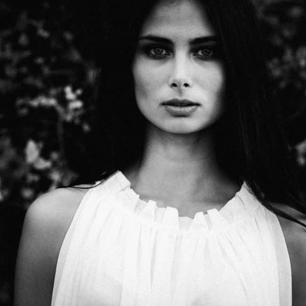 A black and white portrait captures a person with long dark hair and a serious expression, wearing a sleeveless white top. The backdrop of blurred dark foliage adds an element of mystery, offering variety without revealing any clear clue to the true mood behind the gaze.