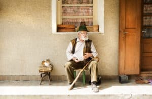 An elderly man in a hat and vest sits on a chair in front of the house, gripping his walking stick like a character from the first murder mystery novel. A cat lounges beside him, while the textured wall and wooden door hint at secrets waiting to be uncovered.