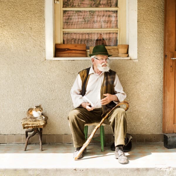 An elderly man in a hat and vest sits on a chair in front of the house, gripping his walking stick like a character from the first murder mystery novel. A cat lounges beside him, while the textured wall and wooden door hint at secrets waiting to be uncovered.