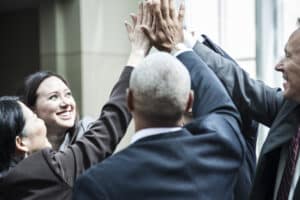 A diverse group of business professionals in suits is gathered, smiling and giving a collective high-five in an office setting. Bright natural light filters through large windows as they celebrate the success of their team building exercises.