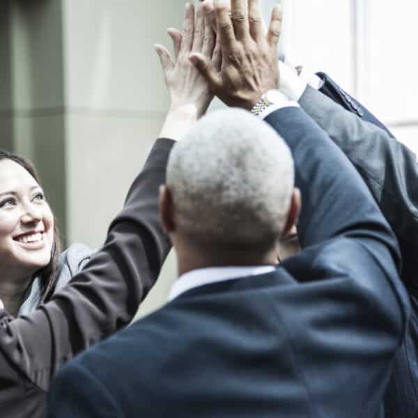 A diverse group of business professionals in suits is gathered, smiling and giving a collective high-five in an office setting. Bright natural light filters through large windows as they celebrate the success of their team building exercises.