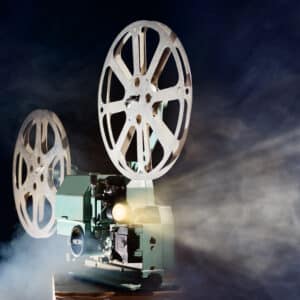 A vintage film projector beams light through cinematic reels, casting a glow amid swirling smoke. The retro device stands against a dark background, evoking the suspense and nostalgia of classic murder mystery movies.