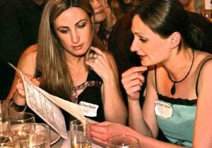 Two women sit at a table in a dimly lit setting, intently examining an interactive seating chart in one womans hand. Glasses are scattered on the table, and both wear name tags, engaging deeply as if planning their next visit to the Apollo Theaters interactive theater experience.