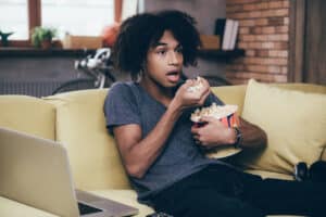 A person with curly hair sits on a couch, looking surprised while eating popcorn and watching one of the best murder mystery TV shows. A laptop rests on their lap, framed by a bicycle in the background, brick walls, and plants on the windowsill.