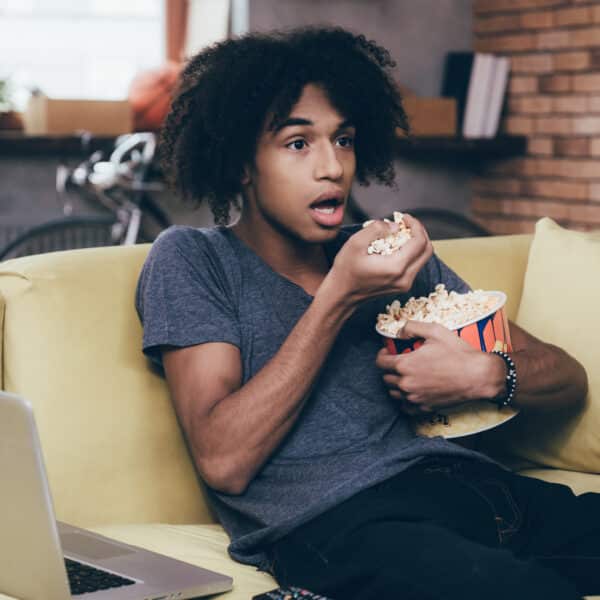 A person with curly hair sits on a couch, looking surprised while eating popcorn and watching one of the best murder mystery TV shows. A laptop rests on their lap, framed by a bicycle in the background, brick walls, and plants on the windowsill.