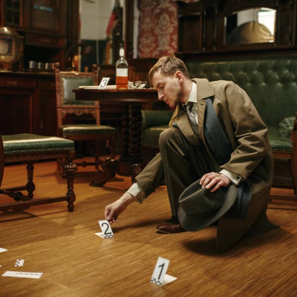 A detective in a trench coat examines evidence markers on the wooden floor of a vintage room. Amidst true crime whispers, a green upholstered bench and a round table with a bottle and card hint at mysteries unsolved.
