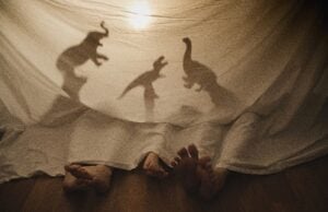 Three pairs of feet peek out from under a sheet, casting shadow puppets of an elephant and two dinosaurs against the fabric. This playful scene evokes the charm of interactive theater, where imagination meets storytelling to create a whimsical atmosphere under soft light.