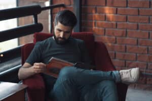 A man with a beard sits in a red chair, engrossed in a crime stories magazine. Hes wearing a dark t-shirt, jeans, and white sneakers. The background showcases a brick wall and black railing, as sunlight streams in from outside.