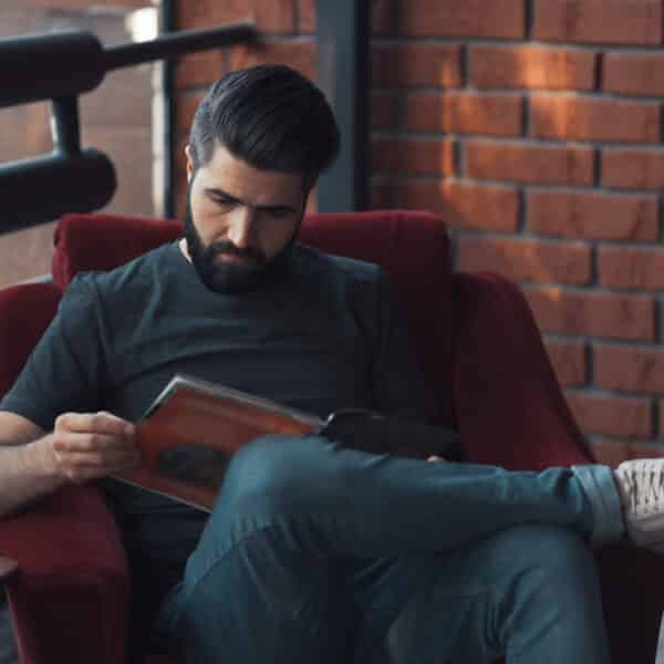 A man with a beard sits in a red chair, engrossed in a crime stories magazine. Hes wearing a dark t-shirt, jeans, and white sneakers. The background showcases a brick wall and black railing, as sunlight streams in from outside.
