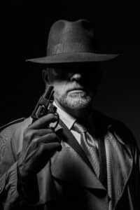 A black-and-white image of a mysterious detective wearing a wide-brimmed hat and trench coat, holding a revolver. His face is partially obscured by shadow, creating an enigmatic atmosphere reminiscent of what makes a good detective in classic noir stories.
