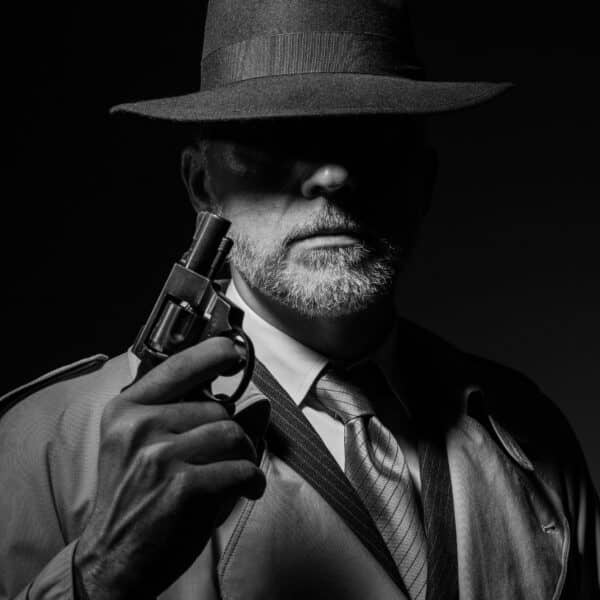 A black-and-white image of a mysterious detective wearing a wide-brimmed hat and trench coat, holding a revolver. His face is partially obscured by shadow, creating an enigmatic atmosphere reminiscent of what makes a good detective in classic noir stories.