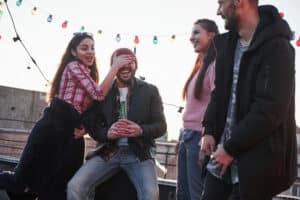 Four friends are having fun at a rooftop gathering. One playfully covers anothers eyes as they hold a beverage, laughter echoing around them. String lights twinkle above, and the sun sets beautifully in the background, highlighting the importance of fun with friends.