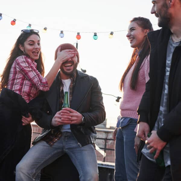 Four friends are having fun at a rooftop gathering. One playfully covers anothers eyes as they hold a beverage, laughter echoing around them. String lights twinkle above, and the sun sets beautifully in the background, highlighting the importance of fun with friends.