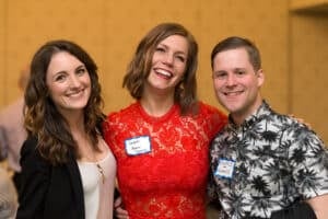 Three people are smiling and posing together at an indoor murder mystery dinner show. The person in the middle is wearing a red lace dress, while the other two are dressed more casually. They are all wearing name tags, adding a touch of intrigue to their identities.