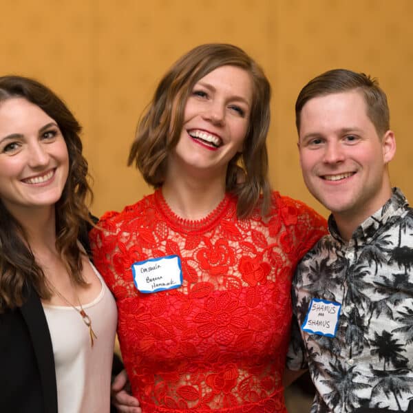Three people are smiling and posing together at an indoor murder mystery dinner show. The person in the middle is wearing a red lace dress, while the other two are dressed more casually. They are all wearing name tags, adding a touch of intrigue to their identities.