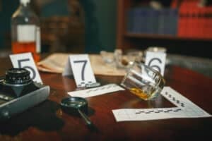 A murder mystery dinner show unfolds on the table, with a fallen whiskey glass beside numbered evidence markers. Nearby, a camera and magnifying glass stand ready. In the blurred background, shelves and scattered papers hint at the secrets waiting to be uncovered.