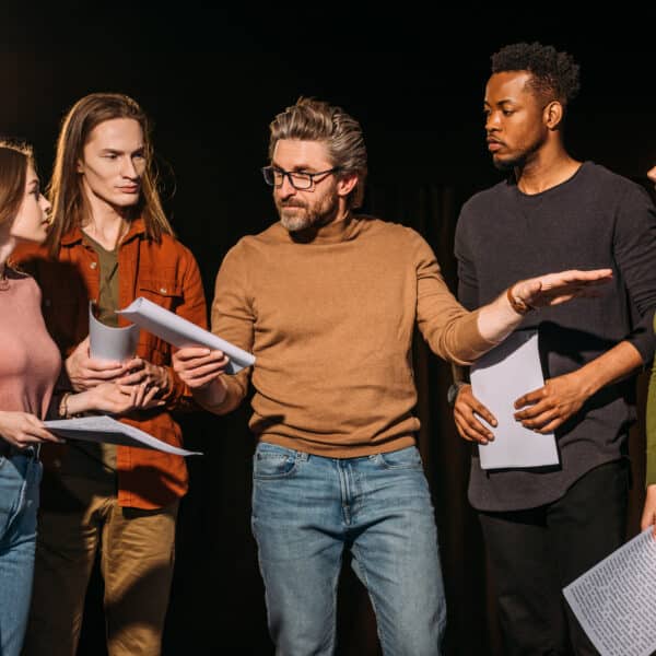 A group of five people stands together under stage lights, holding scripts. One person in the center, wearing glasses, gestures while speaking. The others listen attentively, preparing for their upcoming Murder Mystery Dinner Show performance.