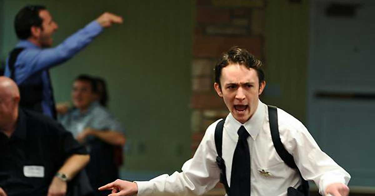 A man in a white dress shirt and black tie dramatically gesturing and speaking in an intense manner during a live performance, with other attendees in the background.
