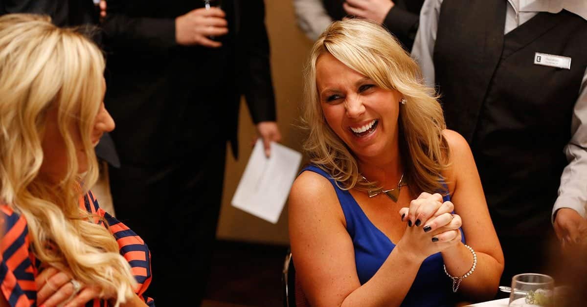 A blonde woman in a blue dress laughing at a dinner table while an event staff member in a black vest stands nearby holding a paper.