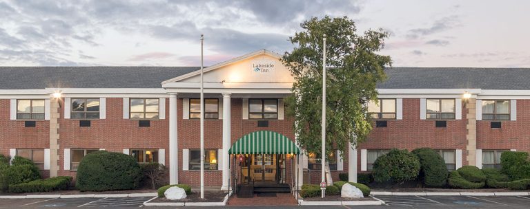 Exterior view of a two-story brick building with a central entrance sheltered by a green canopy, surrounded by well-maintained shrubs. The sky above Norwood is partly cloudy, creating a serene atmosphere.