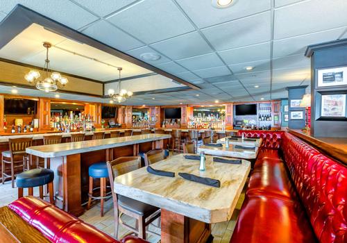A spacious restaurant interior in Norwood features red leather booth seating and wooden tables with black napkins. The bar area with a variety of bottles is visible in the background. Warm lighting from chandeliers creates a cozy atmosphere reminiscent of classic Boston charm.