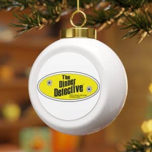 A delicate Christmas Ball Ornament hangs from a tree branch, featuring a yellow oval label reading The Dinner Detective and two bullet hole graphics. The softly blurred background is filled with warm, out-of-focus colors.