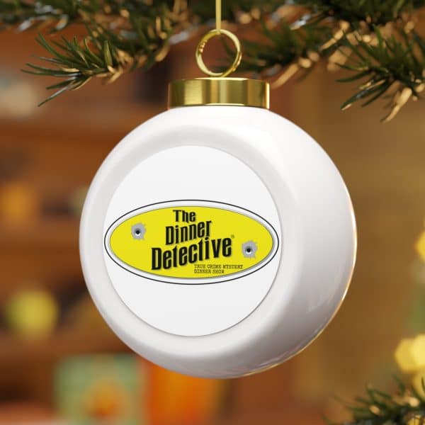 A delicate Christmas Ball Ornament hangs from a tree branch, featuring a yellow oval label reading The Dinner Detective and two bullet hole graphics. The softly blurred background is filled with warm, out-of-focus colors.