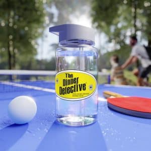 A Tritan Water Bottle labeled The Dinner Detective sits on a blue ping pong table with a white ball and red paddle, against a backdrop of people enjoying nature in a sunlit park.