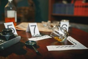 A murder mystery dinner show unfolds on the table, with a fallen whiskey glass beside numbered evidence markers. Nearby, a camera and magnifying glass stand ready. In the blurred background, shelves and scattered papers hint at the secrets waiting to be uncovered.