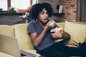 A person with curly hair sits on a couch, looking surprised while eating popcorn and watching one of the best murder mystery TV shows. A laptop rests on their lap, framed by a bicycle in the background, brick walls, and plants on the windowsill.