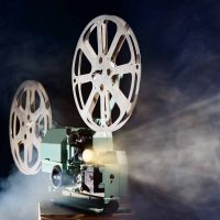 A vintage film projector beams light through cinematic reels, casting a glow amid swirling smoke. The retro device stands against a dark background, evoking the suspense and nostalgia of classic murder mystery movies.