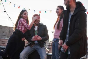 Four friends are having fun at a rooftop gathering. One playfully covers anothers eyes as they hold a beverage, laughter echoing around them. String lights twinkle above, and the sun sets beautifully in the background, highlighting the importance of fun with friends.